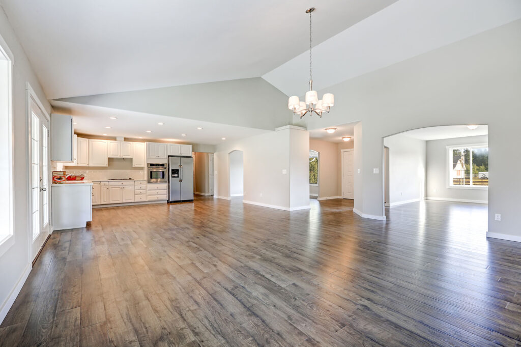 Spacious rambler home interior with vaulted ceiling over glossy luxury vinyl floor. Empty light filled dining or living space adjacent to new white kitchen room features pale grey walls.